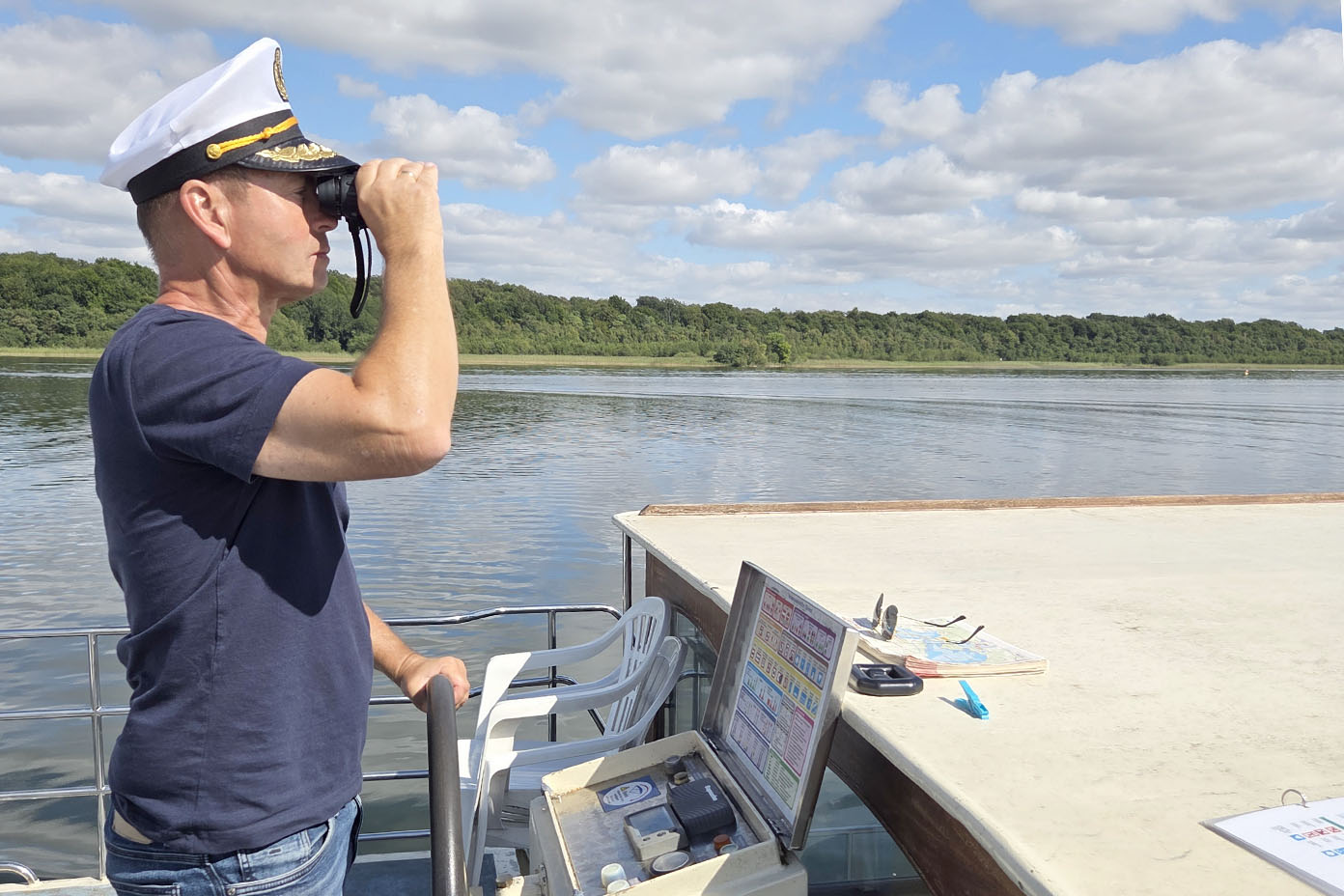 Eine Person mit Kapitänsmütze schaut durch ein Fernglas auf einem Boot, während sie an einem Gewässer mit bewaldetem Ufer steht.