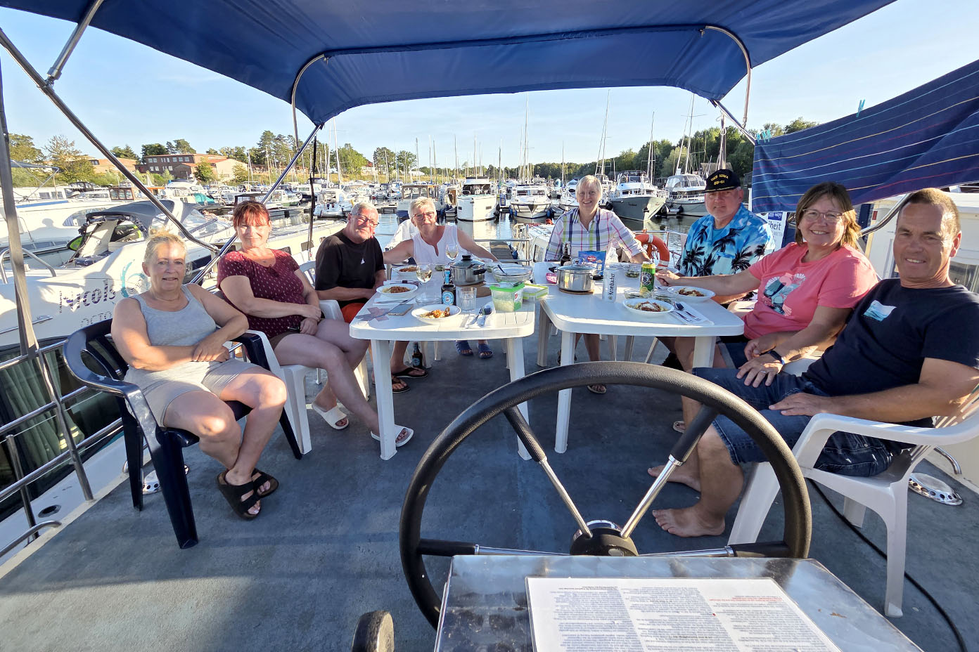 Eine Gruppe von acht Erwachsenen sitzt entspannt auf einem Boot unter einem blauen Sonnendach und genießt gemeinsam eine Mahlzeit mit Blick auf einen Hafen voller Boote.