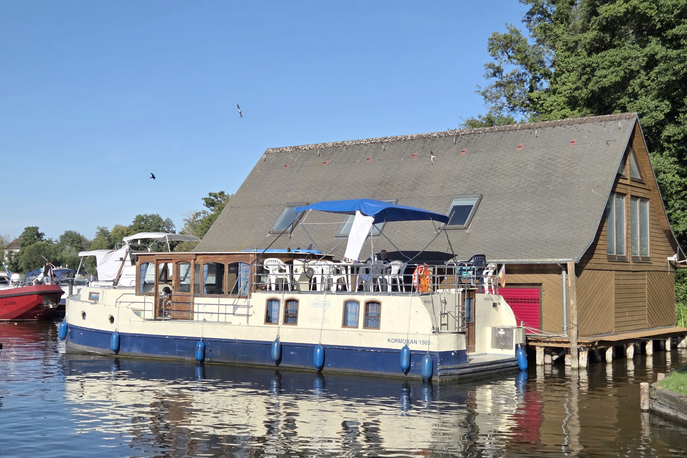 Ein großes Hausboot mit blauer Markise liegt an einem Steg neben einem hölzernen Gebäude mit roter Tür und spitzem Dach an einem ruhigen Gewässer unter klarem Himmel.