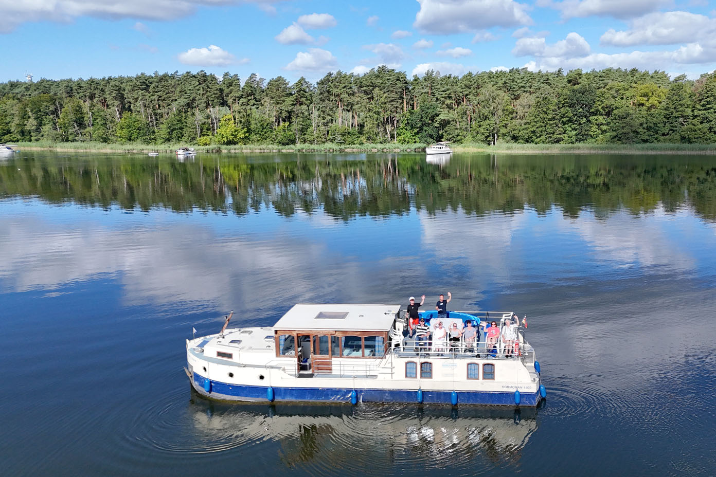 Ein Boot mit mehreren Personen an Deck fährt auf einem ruhigen See, umgeben von Wald und unter einem teilweise bewölkten Himmel.