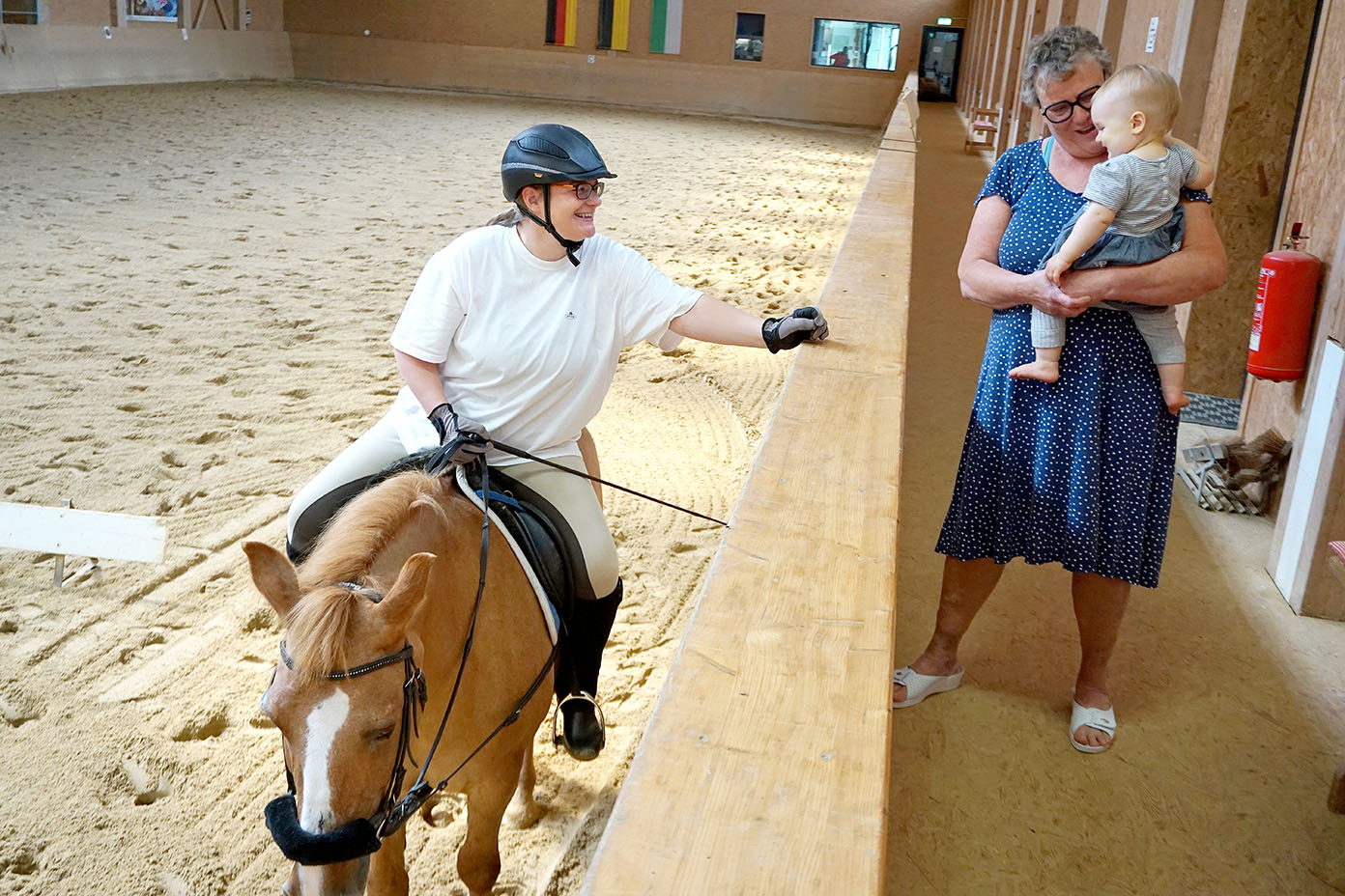 Eine Person in Reitkleidung sitzt auf einem Pferd in einer Reithalle und spricht mit einer anderen Person, die ein Kleinkind im Arm hält.