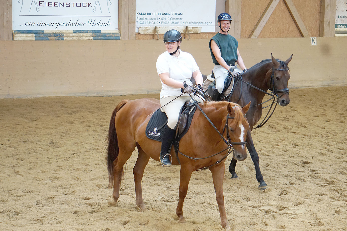 Zwei Personen mit Reithelmen reiten in einer Halle auf braunen Pferden und lächeln dabei.
