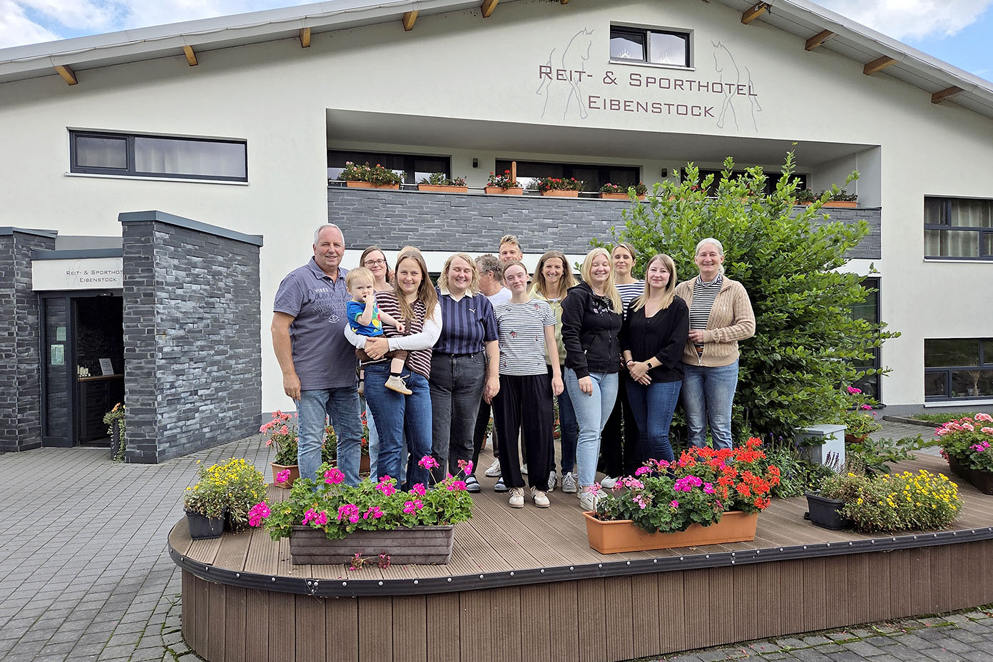 Eine Gruppe von Erwachsenen und einem Kind steht lächelnd vor einem Gebäude mit der Aufschrift Run- & Sporthotel Eibenstock, umgeben von bunten Blumen in Pflanzkästen.