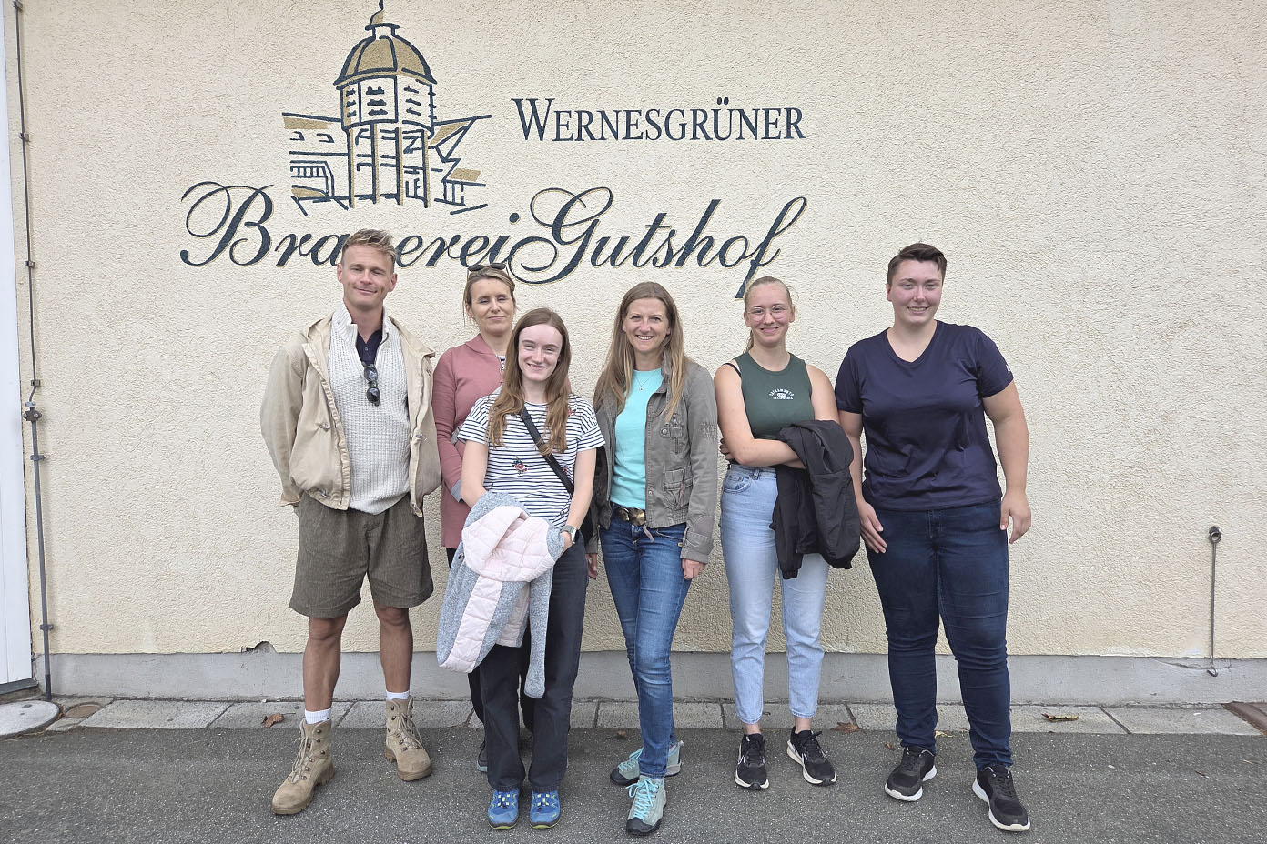 Fünf Personen stehen lächelnd vor einer Wand mit der Aufschrift „Wernesgrüner Brauerei Gutshof“ und tragen legere Kleidung.