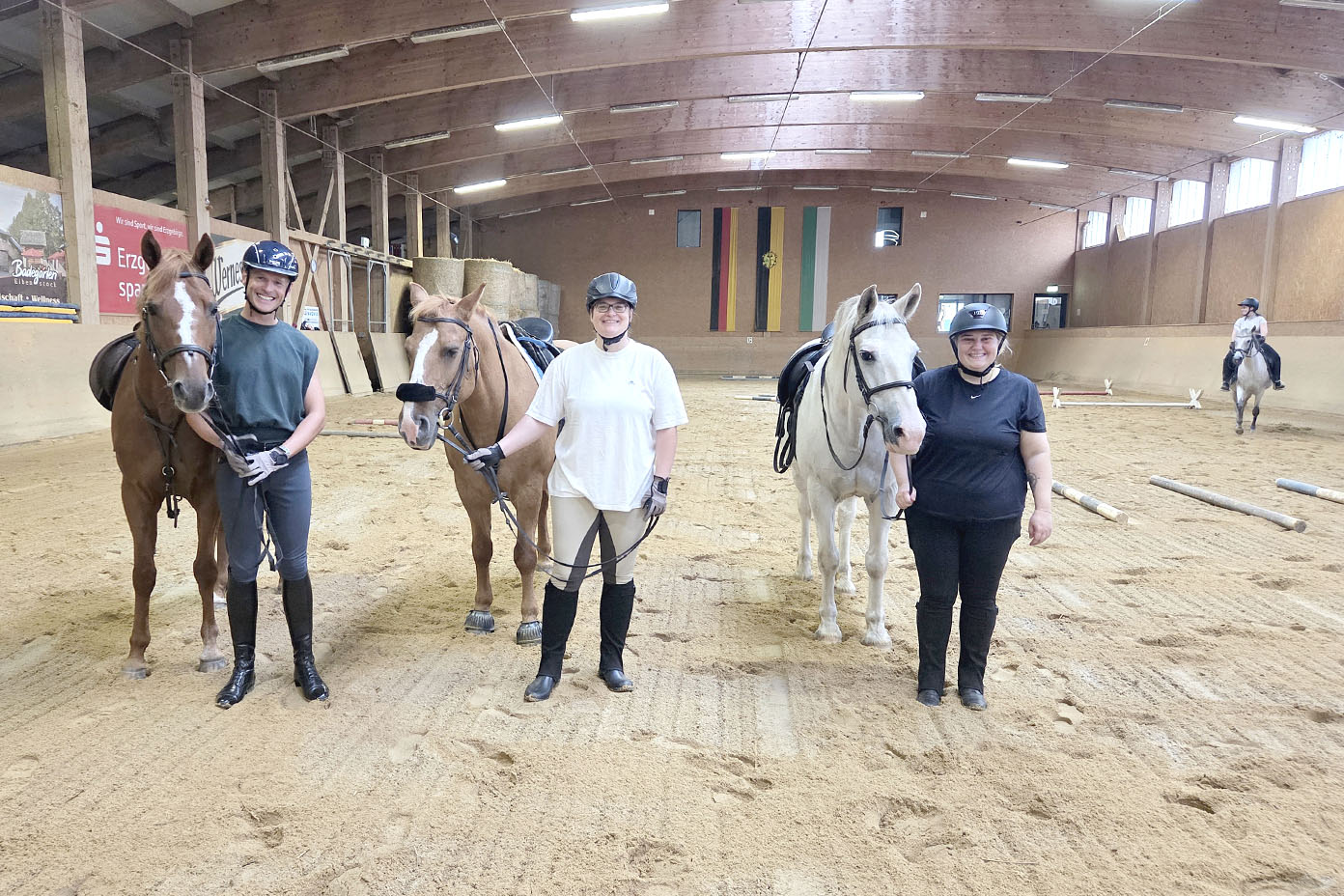 Drei Personen in Reitkleidung stehen in einer Reithalle nebeneinander und halten jeweils ein Pferd an der Hand, während eine weitere Person im Hintergrund auf einem Pferd reitet.