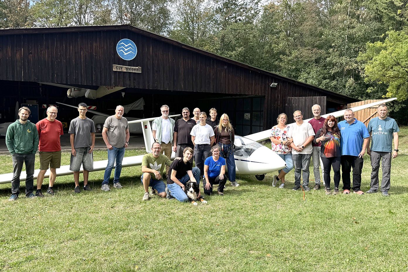 Eine Gruppe von Menschen steht und kniet auf einer Wiese vor einem kleinen Flugzeug und einem Holzgebäude mit einem blauen Schild, umgeben von Bäumen.