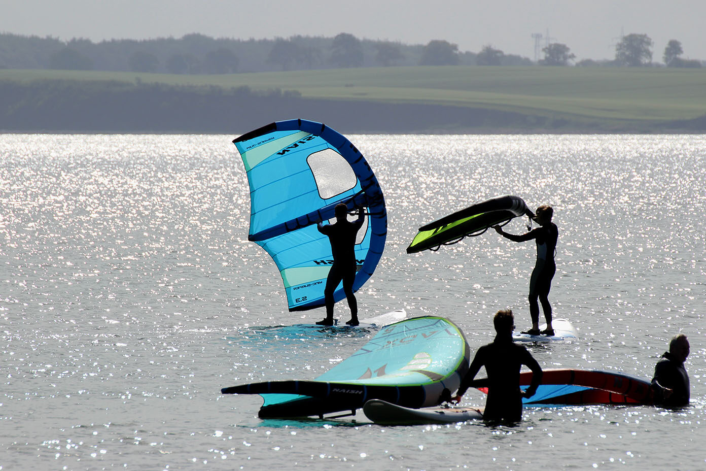 Mehrere Personen stehen auf Wing-Surfboards auf einem ruhigen Gewässer, wobei zwei von ihnen bunte Segel halten, während die Sonne auf dem Wasser glitzert.