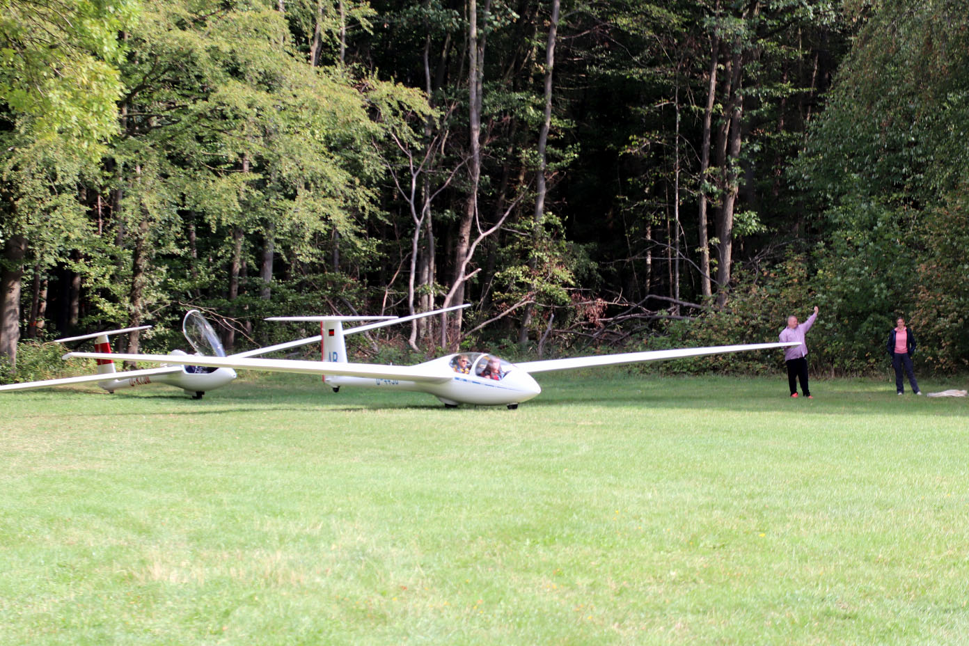 Zwei Segelflugzeuge stehen auf einer Wiese vor einem Wald und zwei Personen stehen in der Nähe der Flugzeuge.