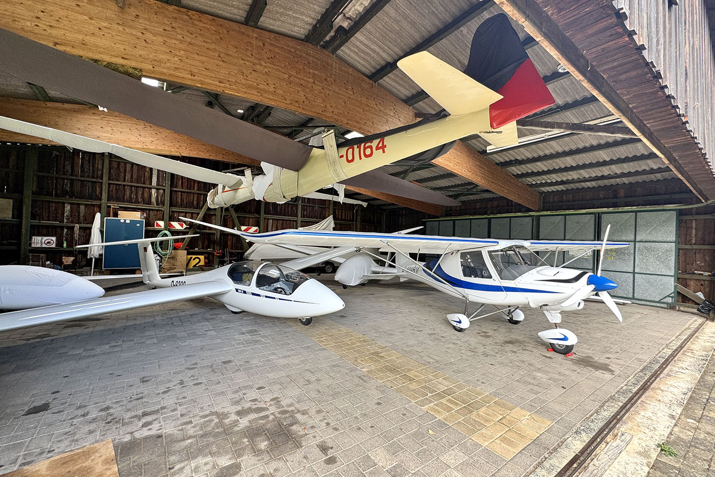 In einem Hangar stehen mehrere kleine Flugzeuge und Segelflugzeuge nebeneinander auf dem Boden.