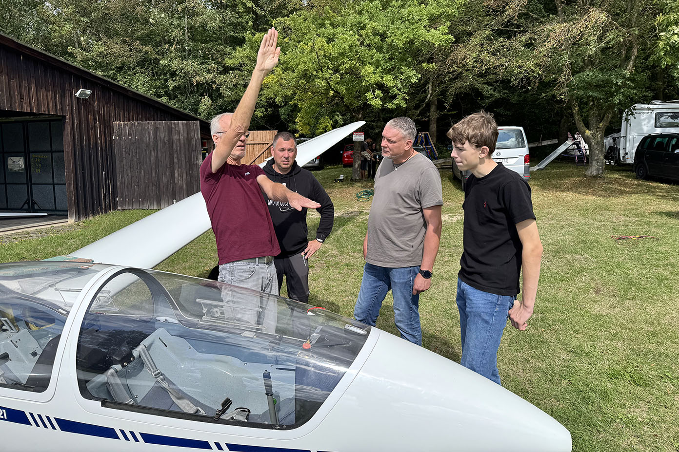 Vier Personen stehen neben einem Segelflugzeug auf einer Wiese, wobei eine Person mit erhobener Hand etwas erklärt.