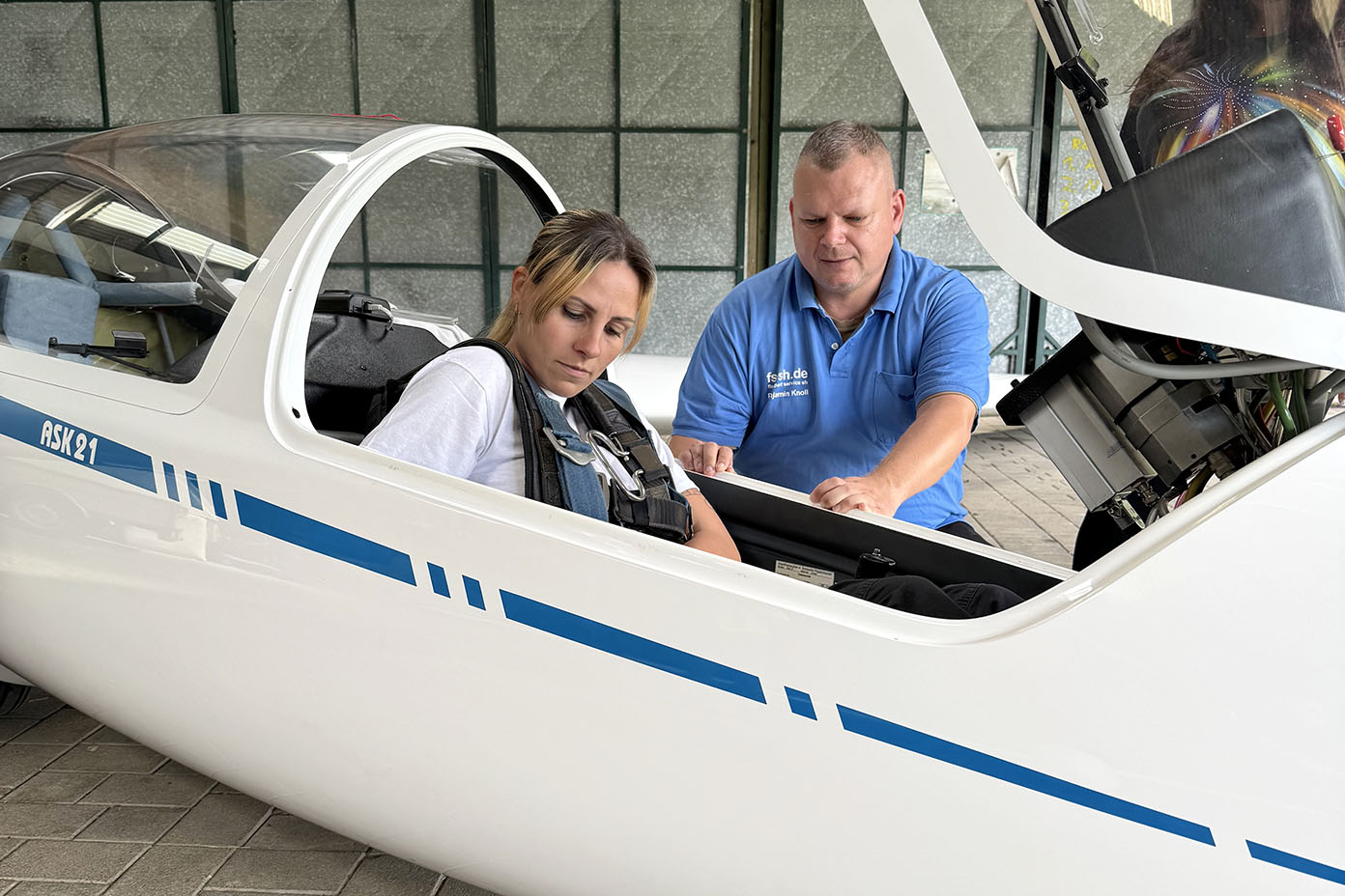 Eine Person mit weißem T-Shirt sitzt in einem Segelflugzeug und eine andere Person in blauem Poloshirt steht daneben und erklärt etwas.