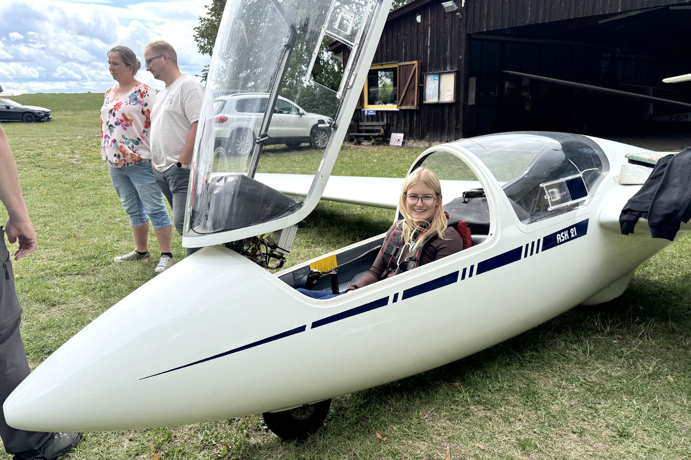 Eine Person mit Brille sitzt lächelnd in einem weißen Segelflugzeug mit geöffnetem Cockpitdach auf einer Wiese, während zwei weitere Personen im Hintergrund stehen.