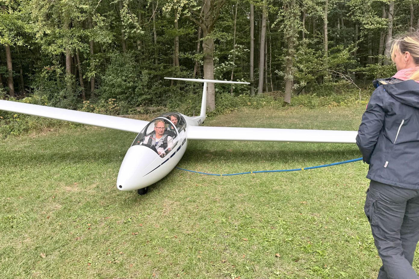 Eine Person sitzt in einem weißen Segelflugzeug auf einer Wiese vor einem Wald und eine weitere Person steht daneben.