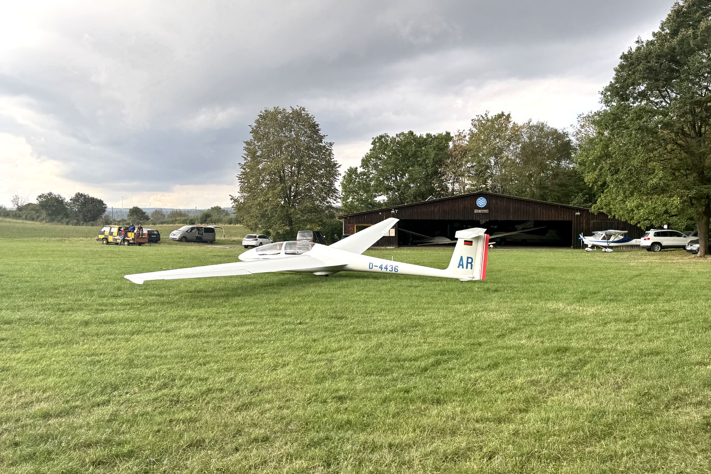 Ein weißes Segelflugzeug steht auf einer großen Wiese vor einem offenen Hangar, umgeben von mehreren Fahrzeugen und Bäumen unter bewölktem Himmel.