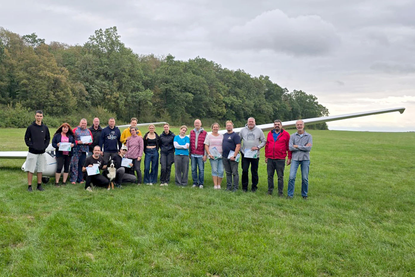 Eine Gruppe von Menschen steht auf einer Wiese vor einem Segelflugzeug mit einem Hund, einige halten Urkunden in der Hand.