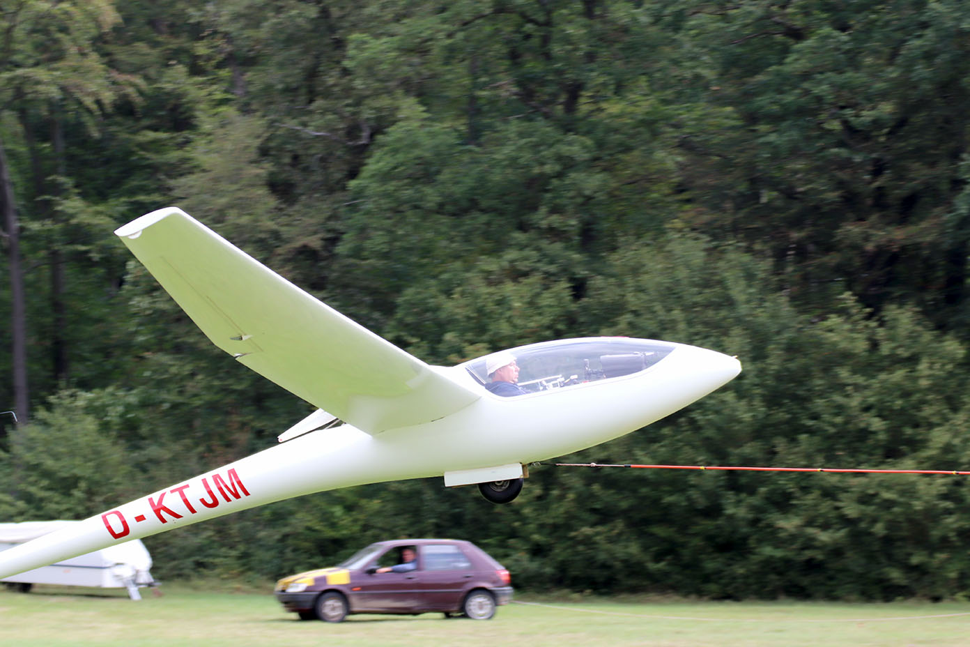 Ein Segelflugzeug wird auf einer Wiese von einem Auto mit einer Zugleine gezogen, im Hintergrund sind Bäume zu sehen.
