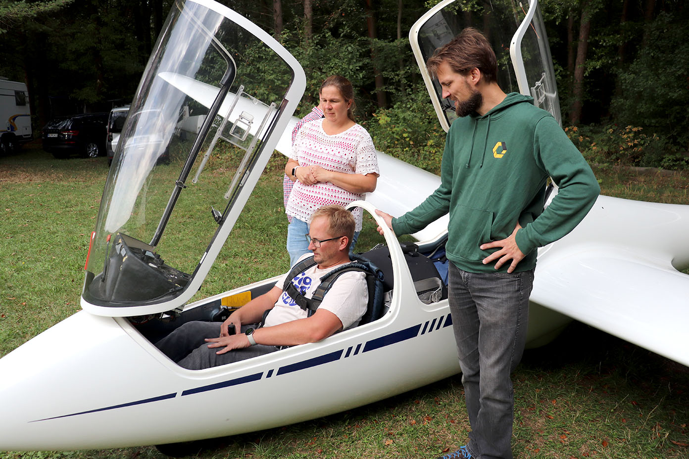 Drei Personen sind an einem Segelflugzeug auf einer Wiese, wobei eine Person im Cockpit sitzt und die anderen beiden daneben stehen und zuschauen.