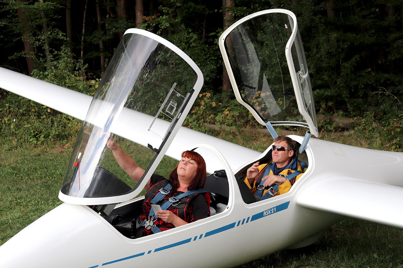 Zwei Personen sitzen in einem Segelflugzeug mit geöffneten transparenten Hauben auf einer Wiese vor einem Wald.