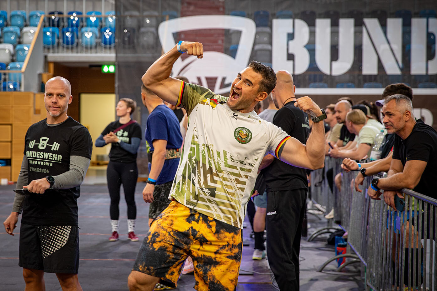 Eine Person in bunten Shorts und einem weißen Sporttrikot zeigt stolz ihre Muskeln in einer Halle, während andere Personen im Hintergrund zuschauen und sich unterhalten.
