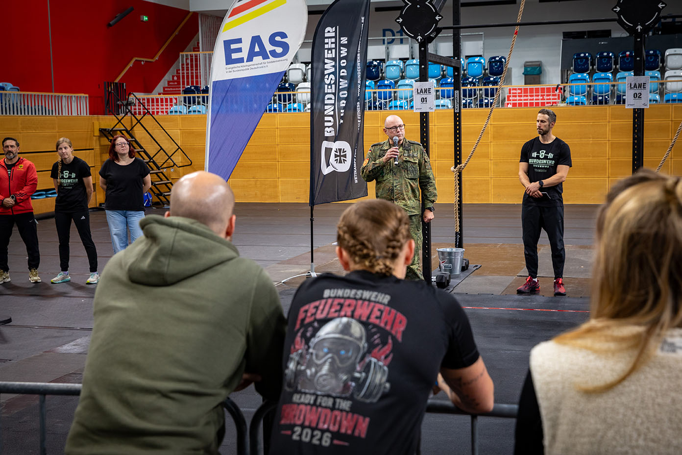 Mehrere Personen stehen in einer Halle vor einer Absperrung, während eine Person in Uniform vor Fahnen und Mikrofonen spricht.