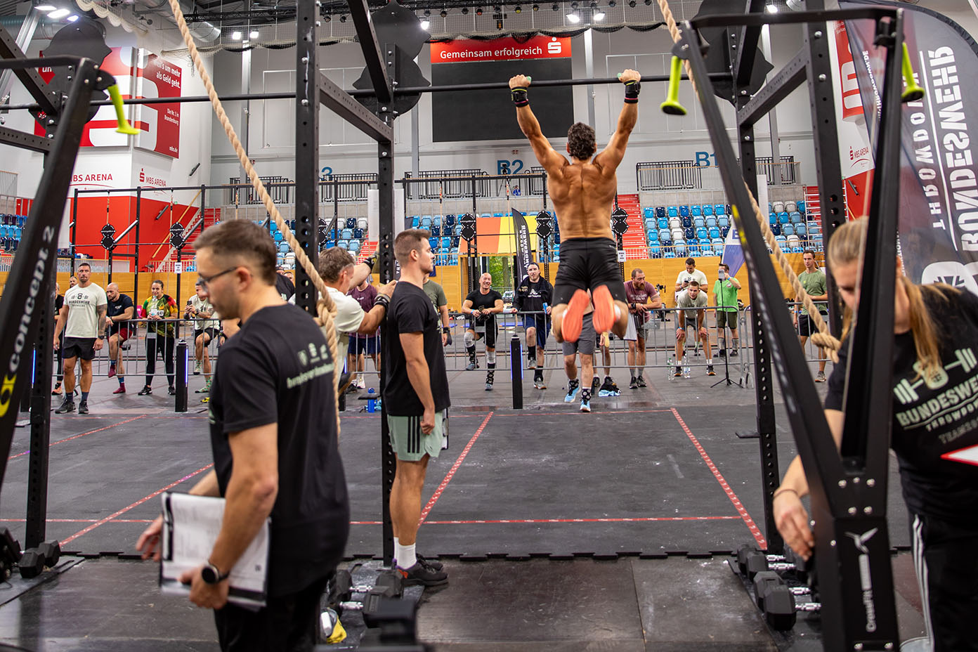 Mehrere Personen nehmen an einem Fitnesswettbewerb in einer Halle teil, wobei eine Person an einer Klimmzugstange hängt und andere zuschauen oder sich vorbereiten.