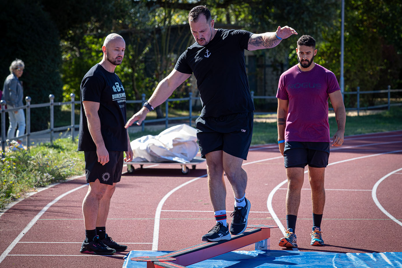 Drei Personen in sportlicher Kleidung stehen auf einer Laufbahn, wobei eine Person auf einem Balancierbalken balanciert und die anderen beiden zuschauen.