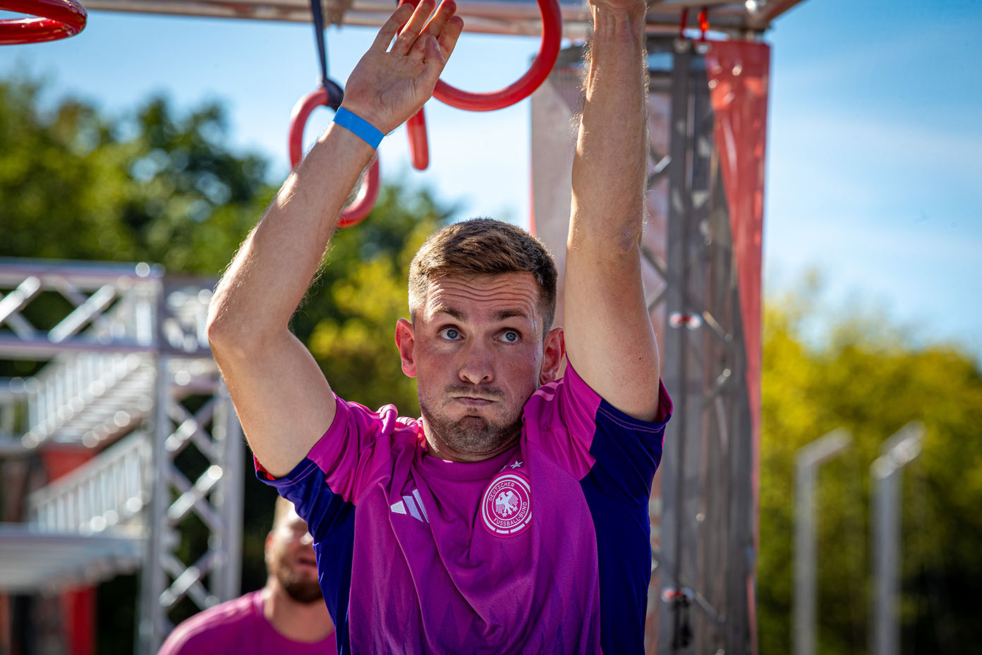 Eine Person in einem lila Sportshirt greift mit beiden Armen nach oben zu roten Turnringen in einer Outdoor-Trainingsumgebung bei sonnigem Wetter.
