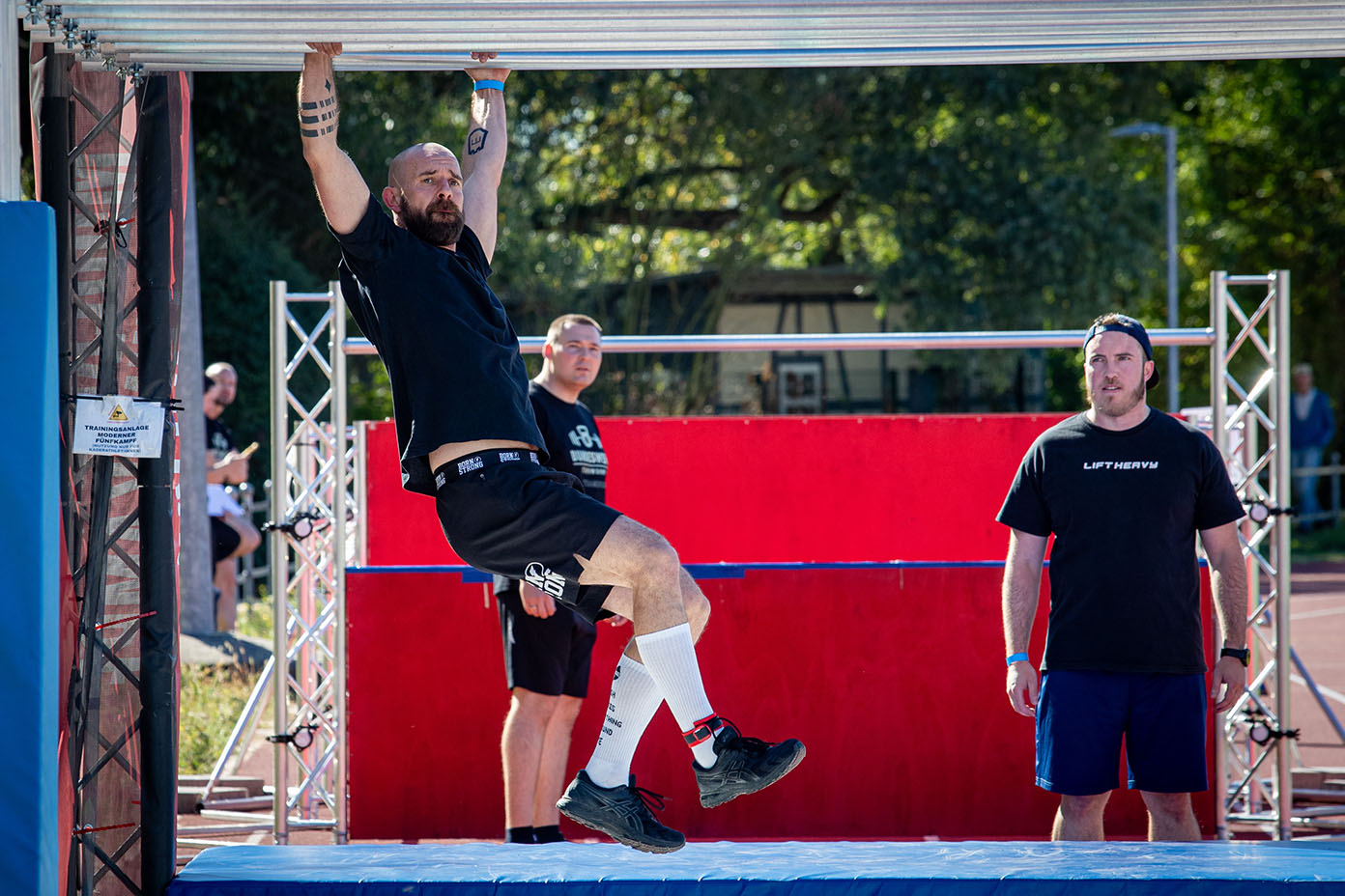 Eine Person in sportlicher Kleidung hängt an einer Stange während zwei weitere Personen danebenstehen und zuschauen, im Hintergrund sind Bäume und eine rote Wand zu sehen.