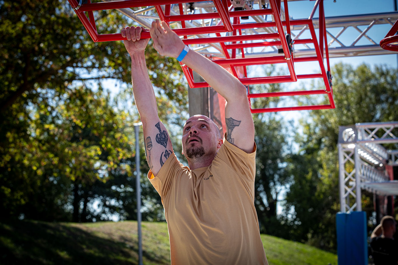 Eine Person mit Tattoos und beigem T-Shirt klettert an einem roten Klettergerüst im Freien bei sonnigem Wetter.