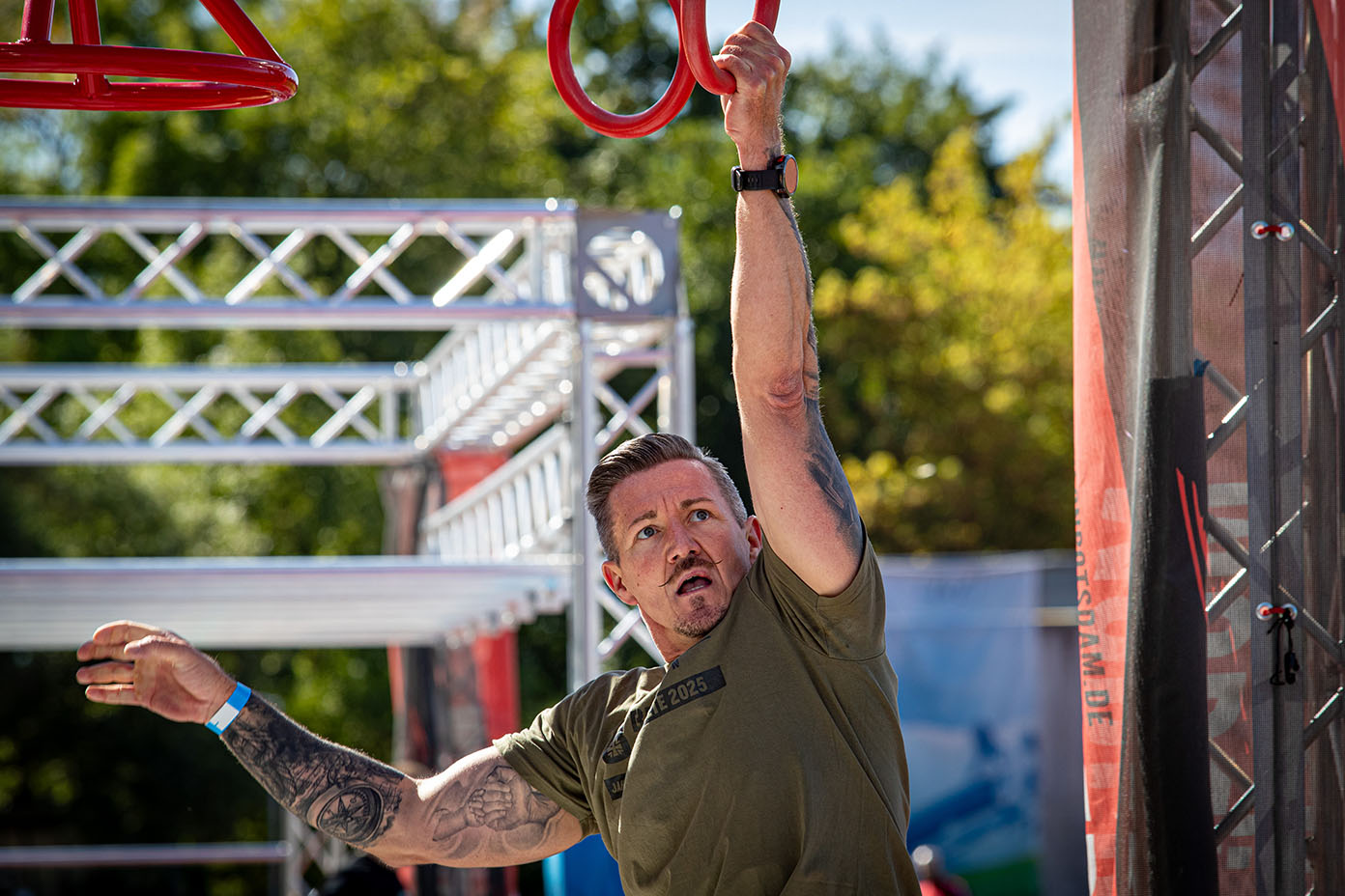 Eine Person mit Tattoos und grünem T-Shirt hängt an einem roten Turnring in einem Outdoor-Hindernisparcours bei sonnigem Wetter.
