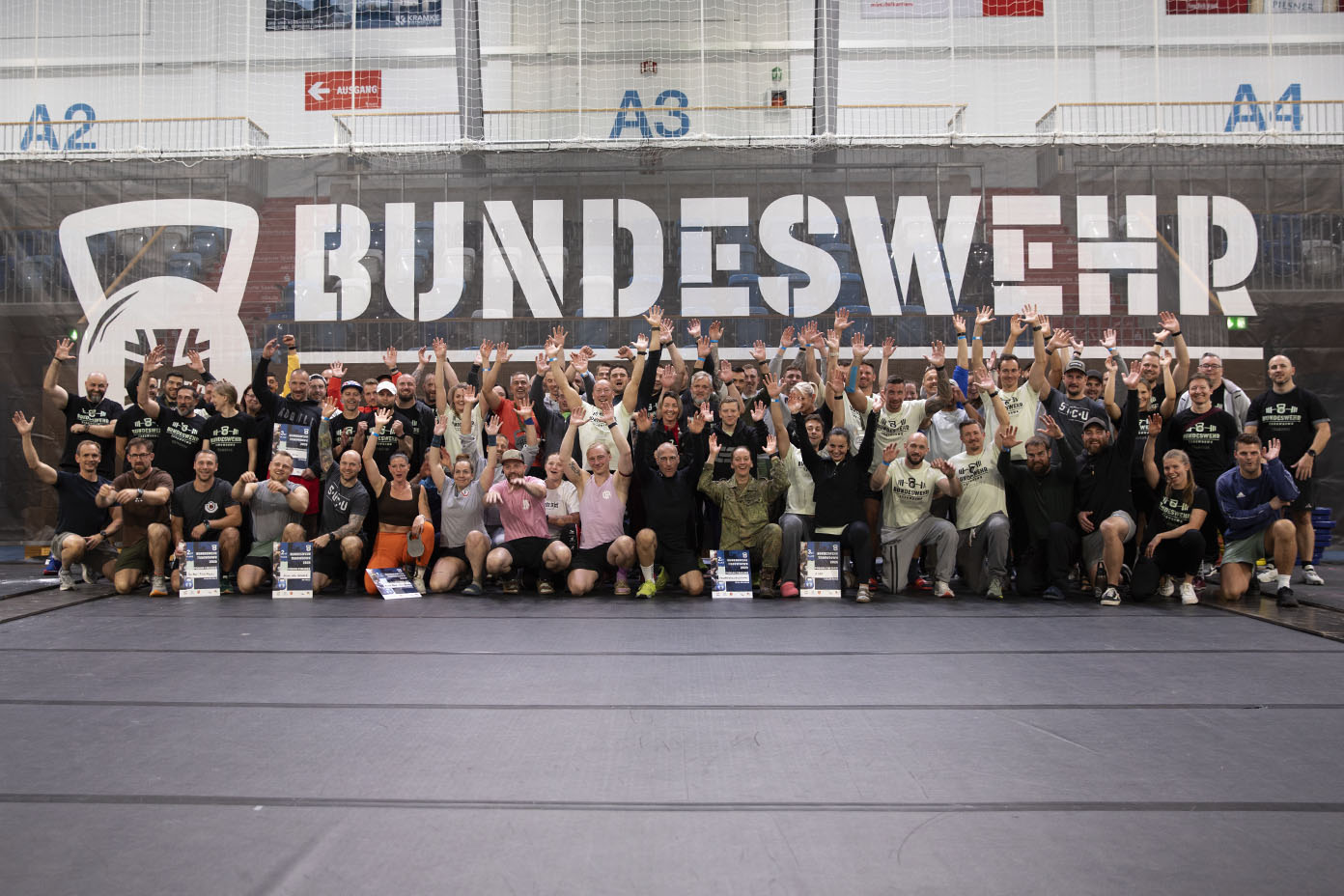 Eine große Gruppe von Menschen in sportlicher Kleidung sitzt und steht vor einem Banner mit der Aufschrift „BUNDESWEHR-THROWDOWN“ und hebt die Hände in die Luft in einer Halle.