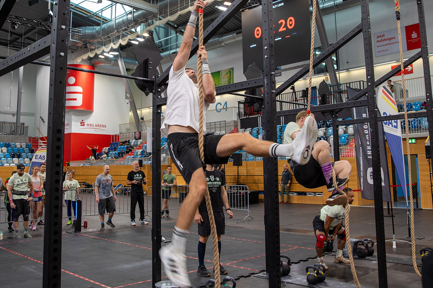 Mehrere Erwachsene in Sportkleidung klettern an Seilen in einer Turnhalle während einer Fitnessveranstaltung mit Zuschauern im Hintergrund.