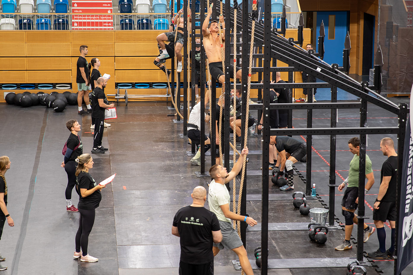 Mehrere Personen trainieren in einer Halle an Kletterstangen und mit Kettlebells, während einige andere Personen mit Klemmbrettern die Übungen beobachten und notieren.