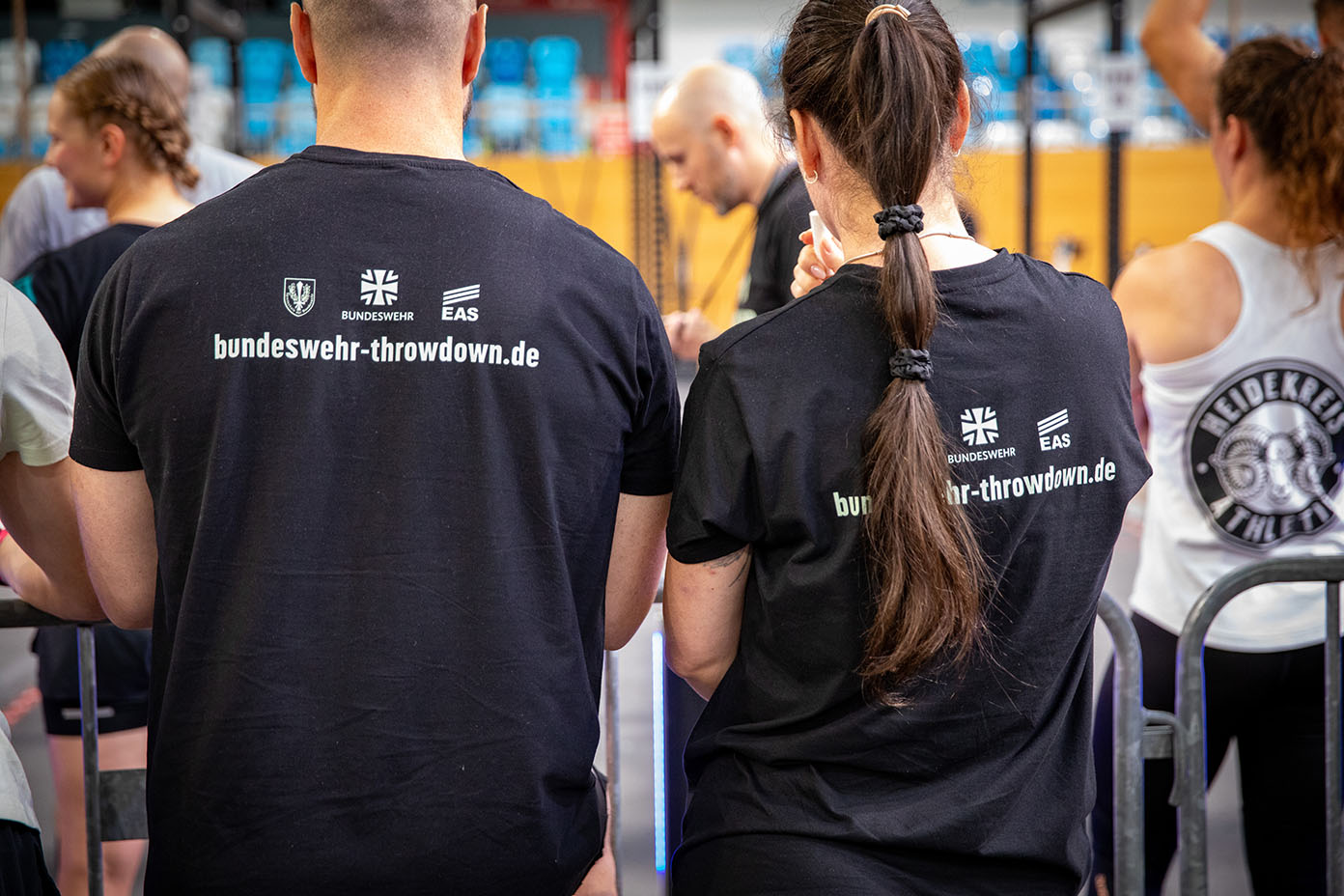 Zwei Personen mit schwarzen T-Shirts, auf denen "bundeswehr-throwdown.de" steht, sitzen in einer Sporthalle mit weiteren Personen im Hintergrund.