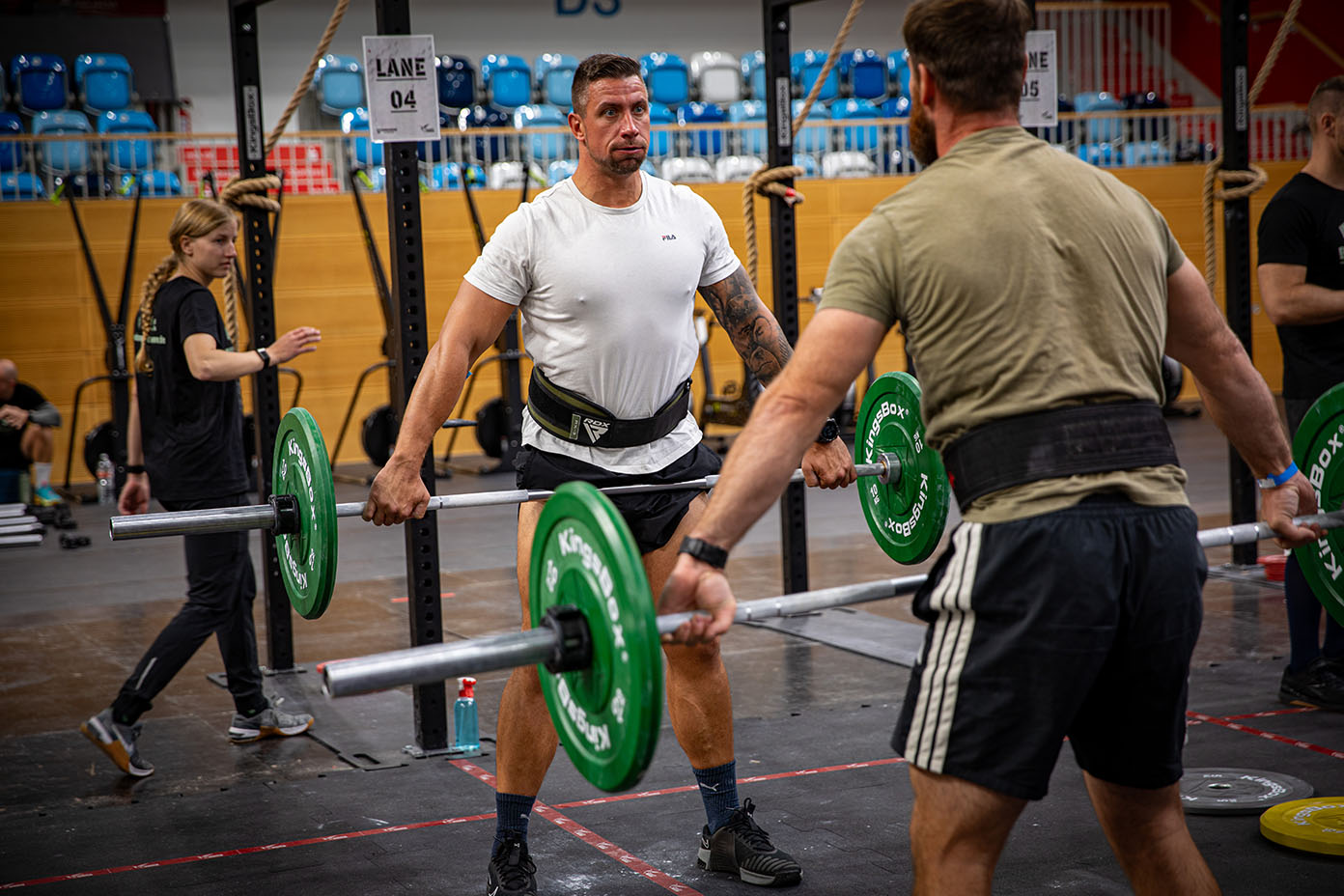 Zwei Personen heben in einer Halle mit Gewichthebergürteln an Langhanteln mit grünen Gewichtsscheiben während eines Trainingswettkampfs.