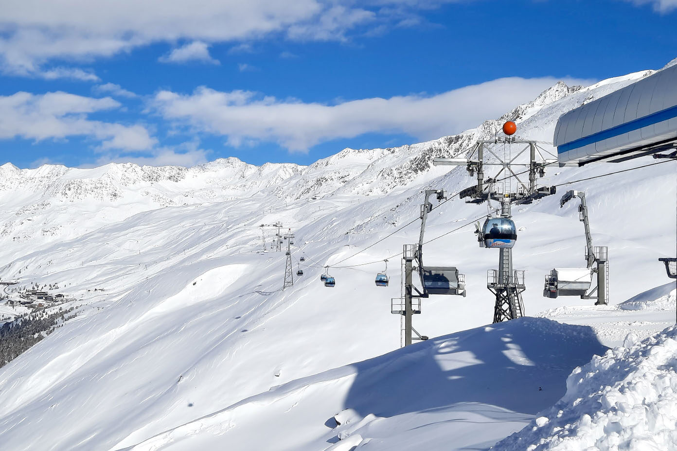 Eine Seilbahn fährt über eine schneebedeckte Berglandschaft unter einem blauen Himmel mit vereinzelten Wolken.