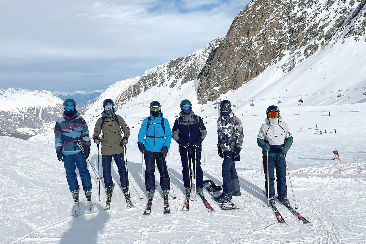 Sechs Personen in bunter Skibekleidung stehen mit Skiern und Snowboard auf einer schneebedeckten Berglandschaft unter einem bewölkten Himmel.