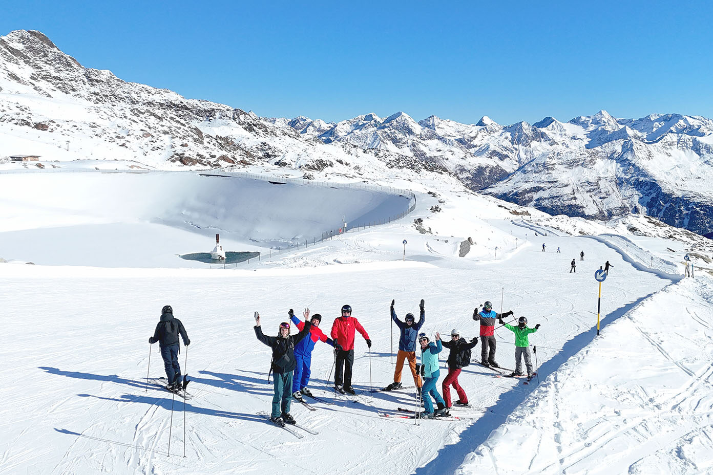 Eine Gruppe von Menschen in bunter Skibekleidung steht auf einer schneebedeckten Piste in den Bergen bei klarem Himmel und hebt die Arme freudig.